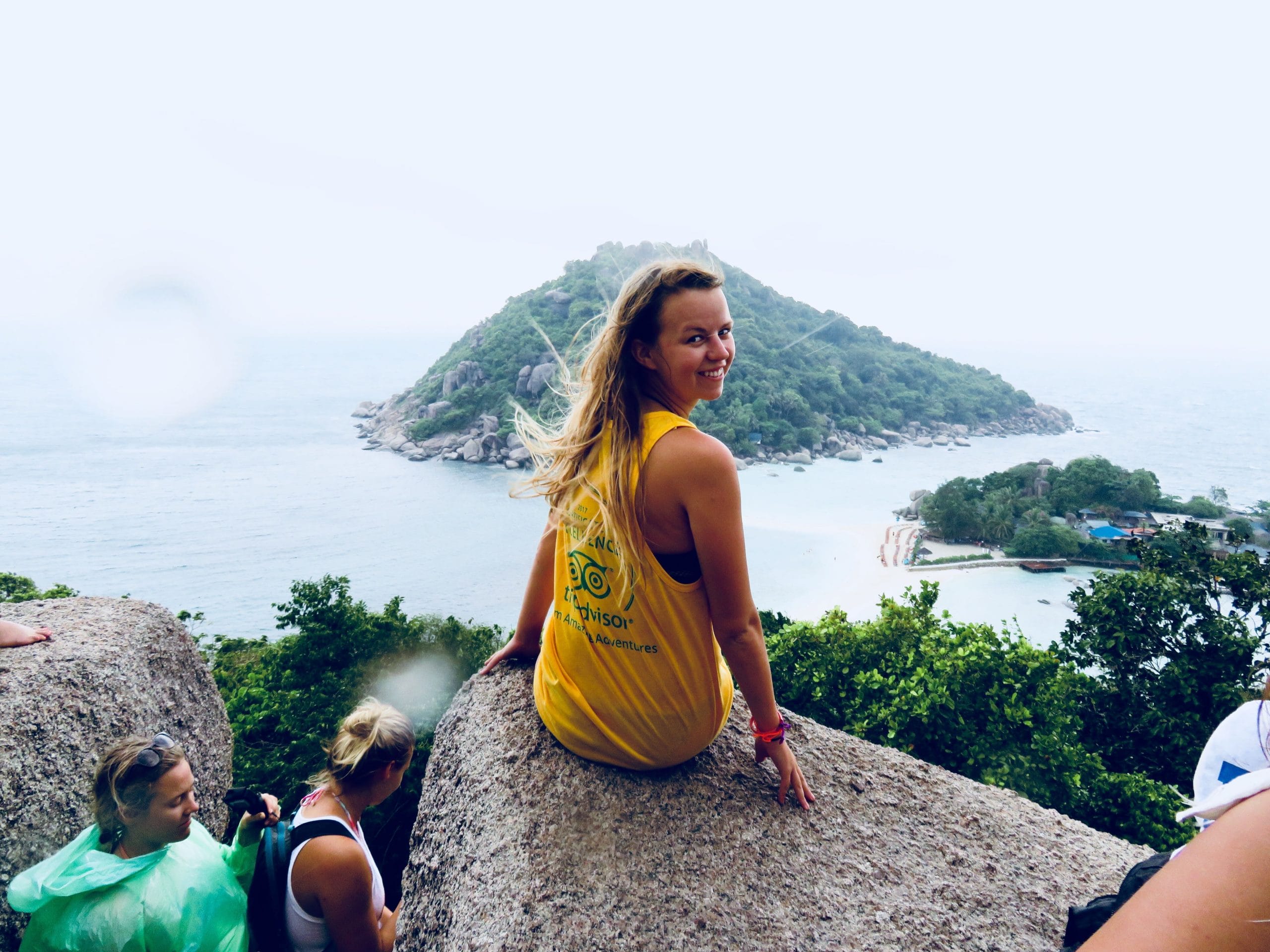 A woman in a yellow tank top enjoys her Lifty Life, smiling at the camera as she sits on a rock with an island and the ocean behind her; other people are nearby.