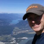 A person wearing a cap enjoys the Lifty Life, taking a selfie on a mountain with rivers, fields, and distant mountains stretching under a clear blue sky.