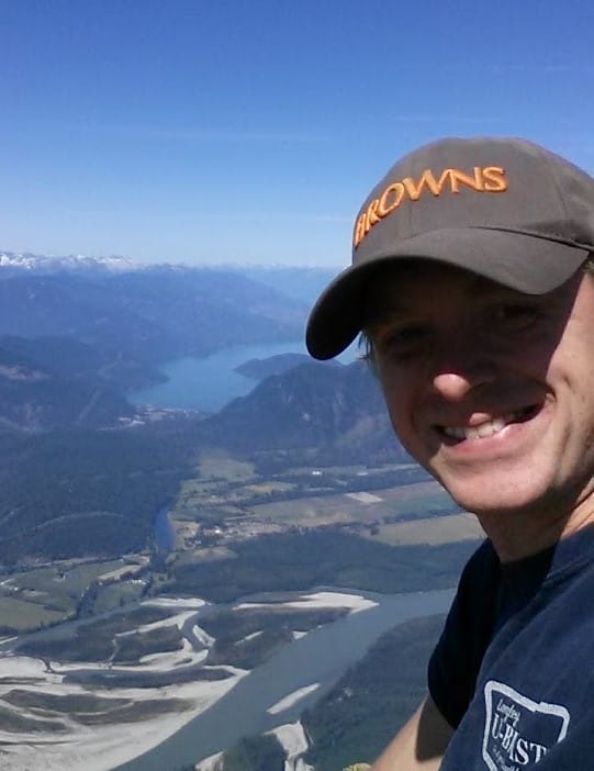A person wearing a cap enjoys the Lifty Life, taking a selfie on a mountain with rivers, fields, and distant mountains stretching under a clear blue sky.