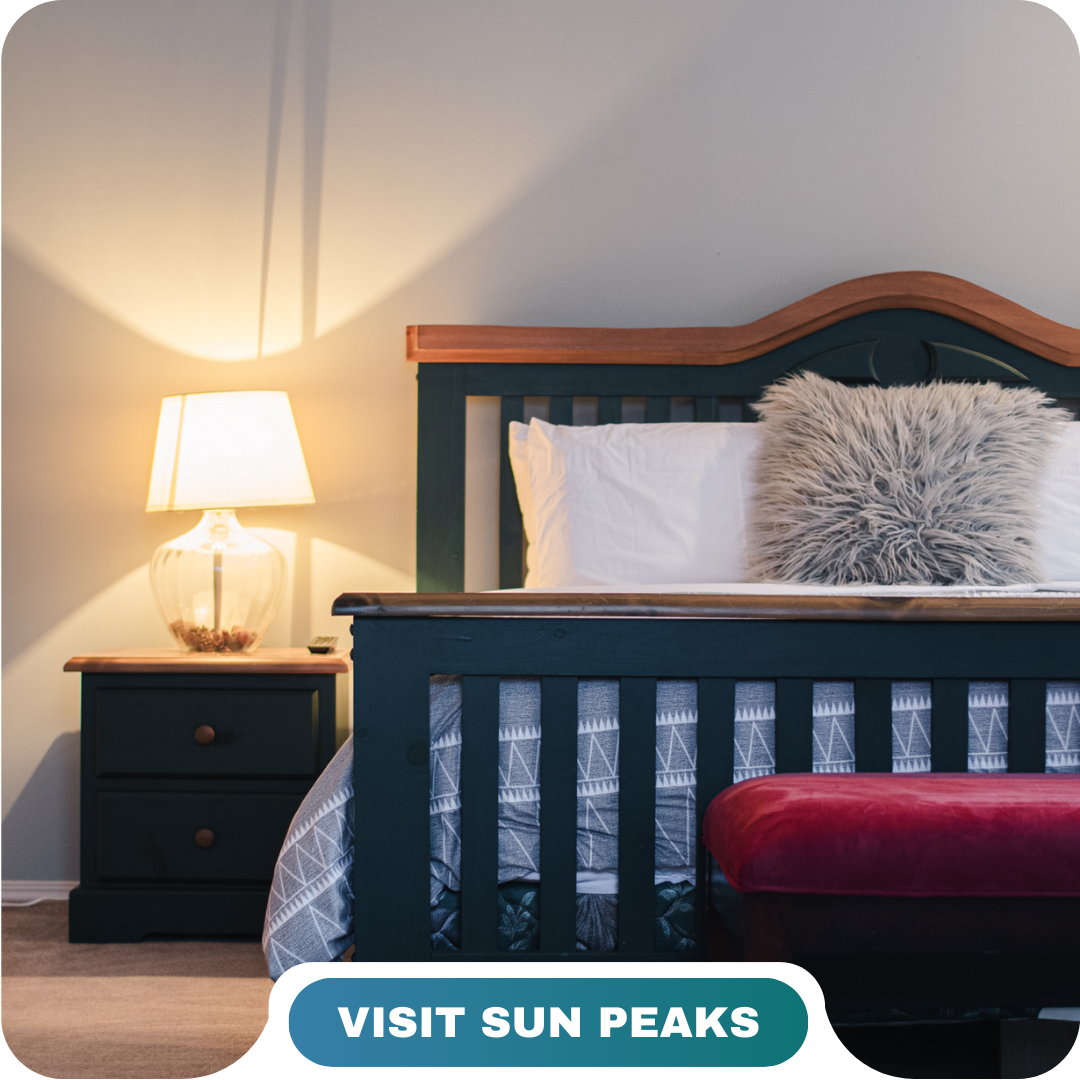 A neatly made bed with a gray fur pillow, wooden headboard, bedside table, and lamp. A red bench sits at the foot of the bed. Text reads "Visit Sun Peaks." Learn more about us and discover your perfect getaway.