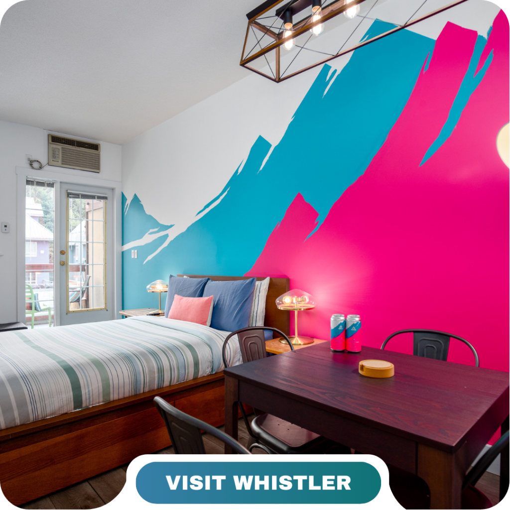 A modern bedroom with a striped bed, wall art of blue and pink mountains, a small dining table, and a "Visit Whistler" label at the bottom—perfect for enjoying Lifty Life comfort.