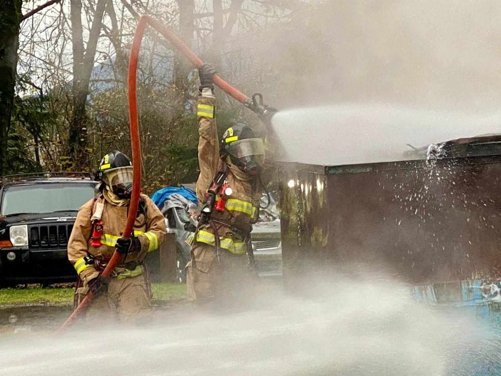 Two firefighters in full gear, part of our dedicated about us team, spray water from hoses onto a smoking dumpster outdoors, with trees and vehicles in the background.