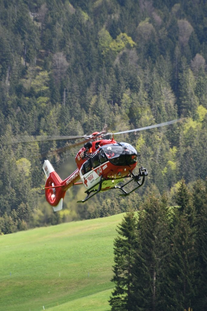 A red rescue helicopter, part of our About Us story, flies low over a grassy field with dense green forest in the background.