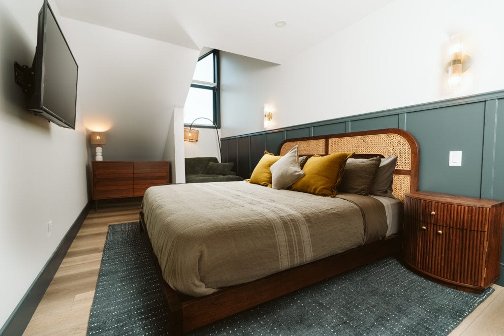 Modern bedroom with a large bed, throw pillows, wooden furniture, wall-mounted TV, and a corner window letting in natural light—perfect for guests seeking comfort through vacation rental management services.