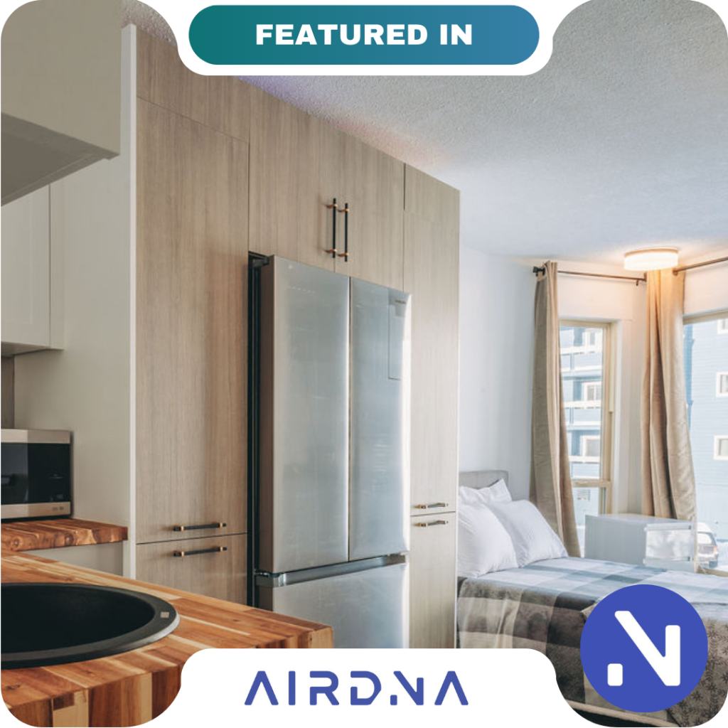 Modern studio apartment with light wood cabinetry, stainless steel refrigerator, and a neatly made bed near large windows. "FEATURED IN AIRDNA PARTNERS" branding is overlaid at the top and bottom.