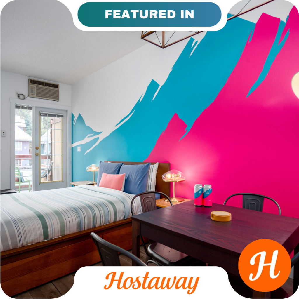 Modern bedroom with a bold blue and pink accent wall, striped bedding, desk with chairs, and glass door leading outside. "Hostaway" and "Featured In" banners highlight trusted partners in the image.
