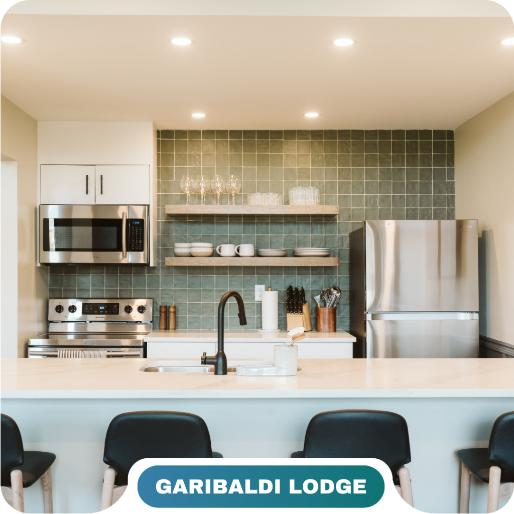 Modern kitchen with stainless steel appliances, open shelves with dishes and glasses, green tile backsplash, and a white countertop with three barstools—perfect for vacation rentals in Whistler.