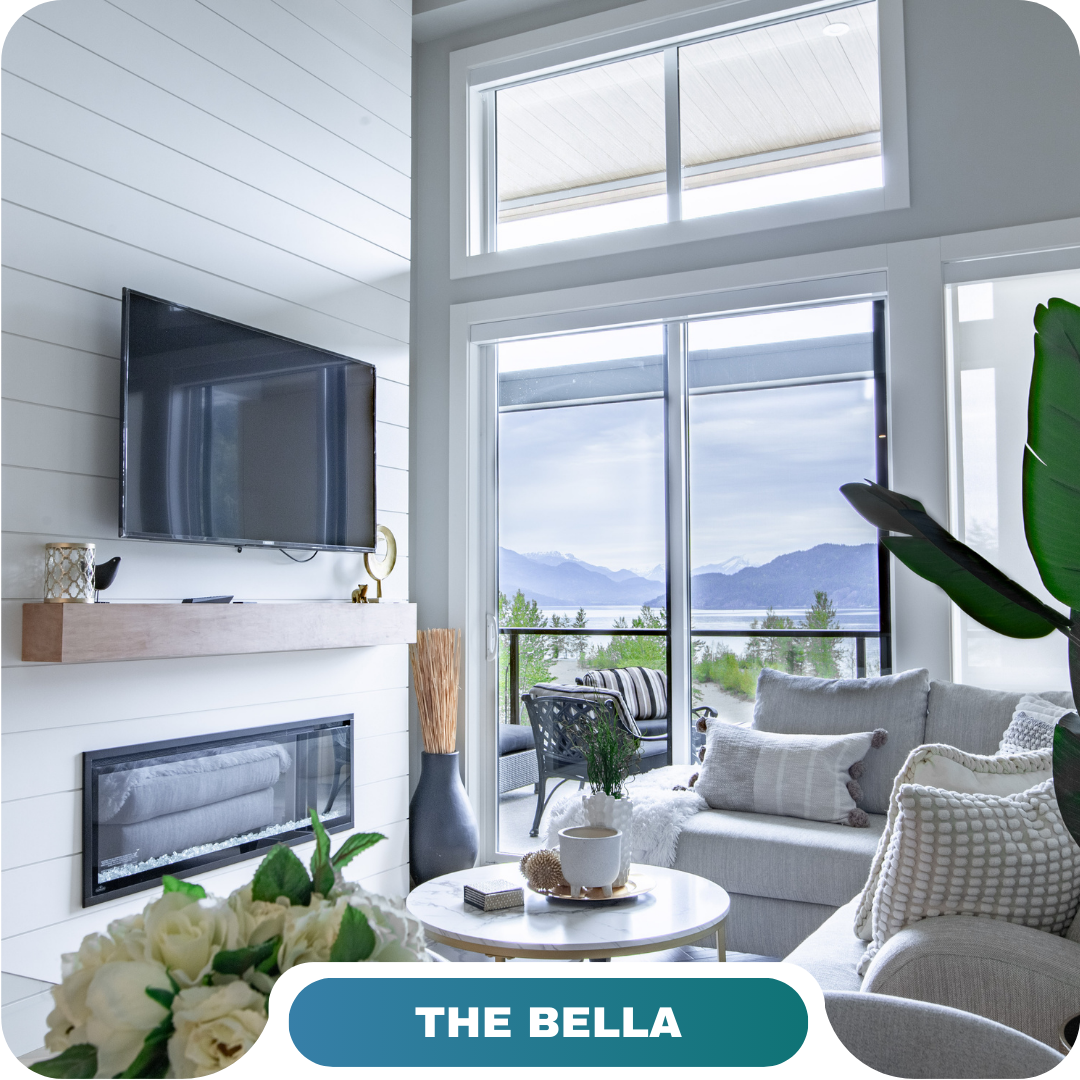 Modern living room with a mounted TV, fireplace, large windows showcasing a mountain view, and neutral-toned furniture—perfect for vacation rentals in Harrison Hot Springs; text reads "The Bella.