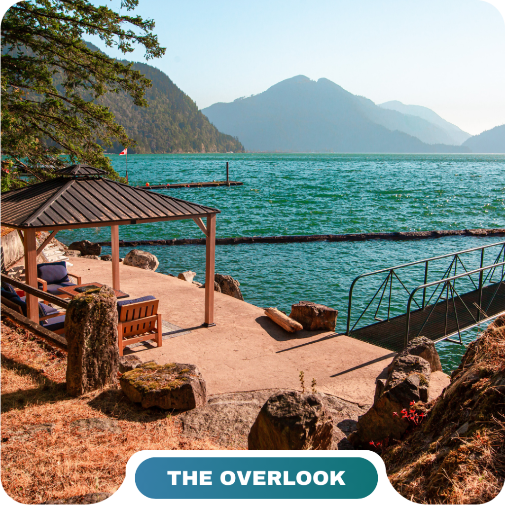 A lakeside gazebo and bench overlook turquoise water, with a dock, mountains, and trees in the background—an inviting scene perfect for vacation rentals in Harrison Hot Springs under a clear sky.