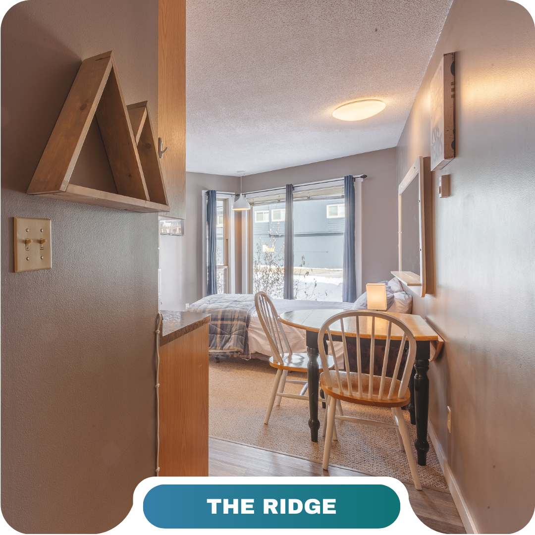 A small, tidy bedroom in one of the vacation rentals in Big White, featuring a desk, two chairs, a cozy bed, large curtained windows, and a wooden shelf shaped like triangles.