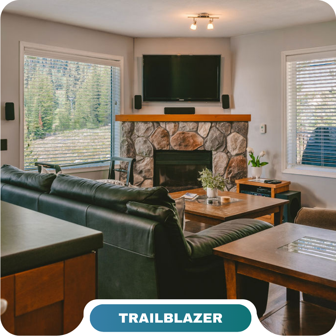 A cozy living room with a stone fireplace, wall-mounted TV, black leather sofa, and large windows showing a forested mountain view—perfect for vacation rentals in Big White.