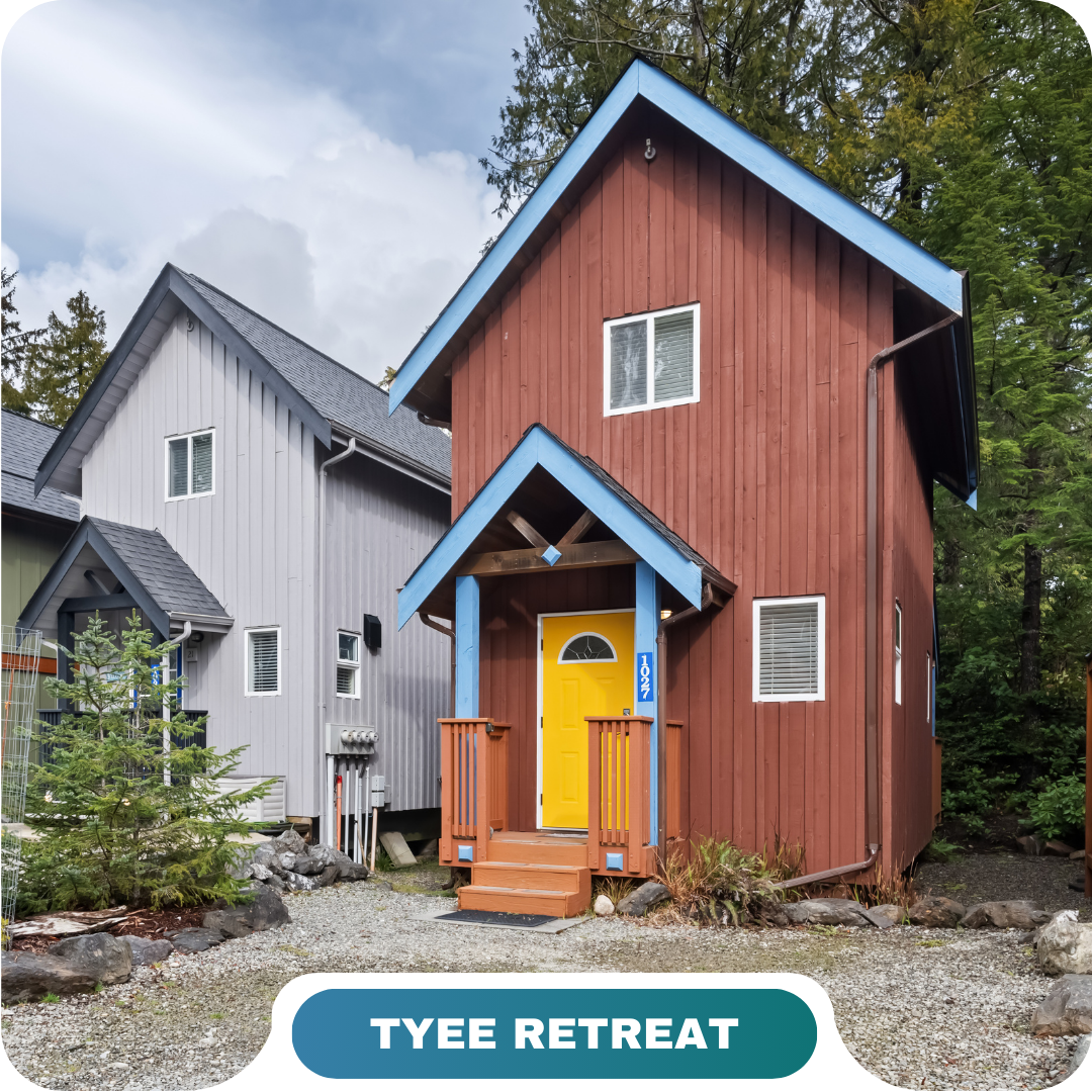 A small brown wooden cabin with a yellow door and blue trim, perfect for vacation rentals in Big White, sits next to a gray house, surrounded by trees and gravel.