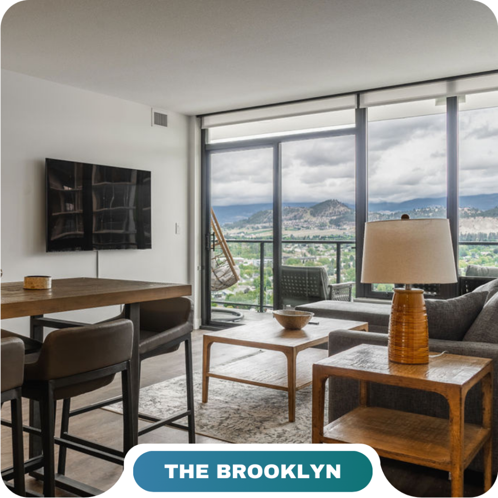 Modern apartment living room with a mounted TV, dining table, and large windows offering a mountain and city view; “The Brooklyn” is perfect for those seeking vacation rentals in Big White.