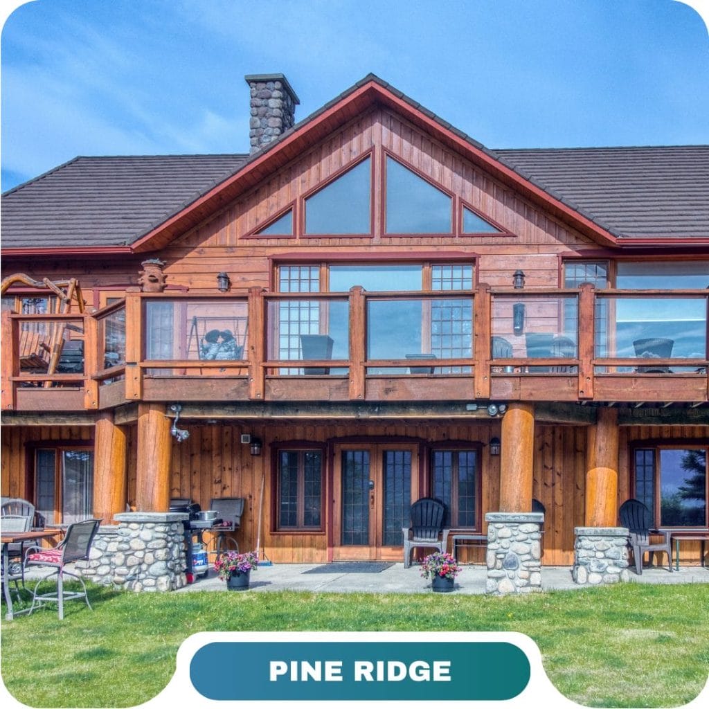 A two-story wooden cabin with large windows, a stone chimney, and a balcony sits on a grassy lawn—ideal for vacation rentals in Invermere & Panorama. The text "PINE RIDGE" appears at the bottom.