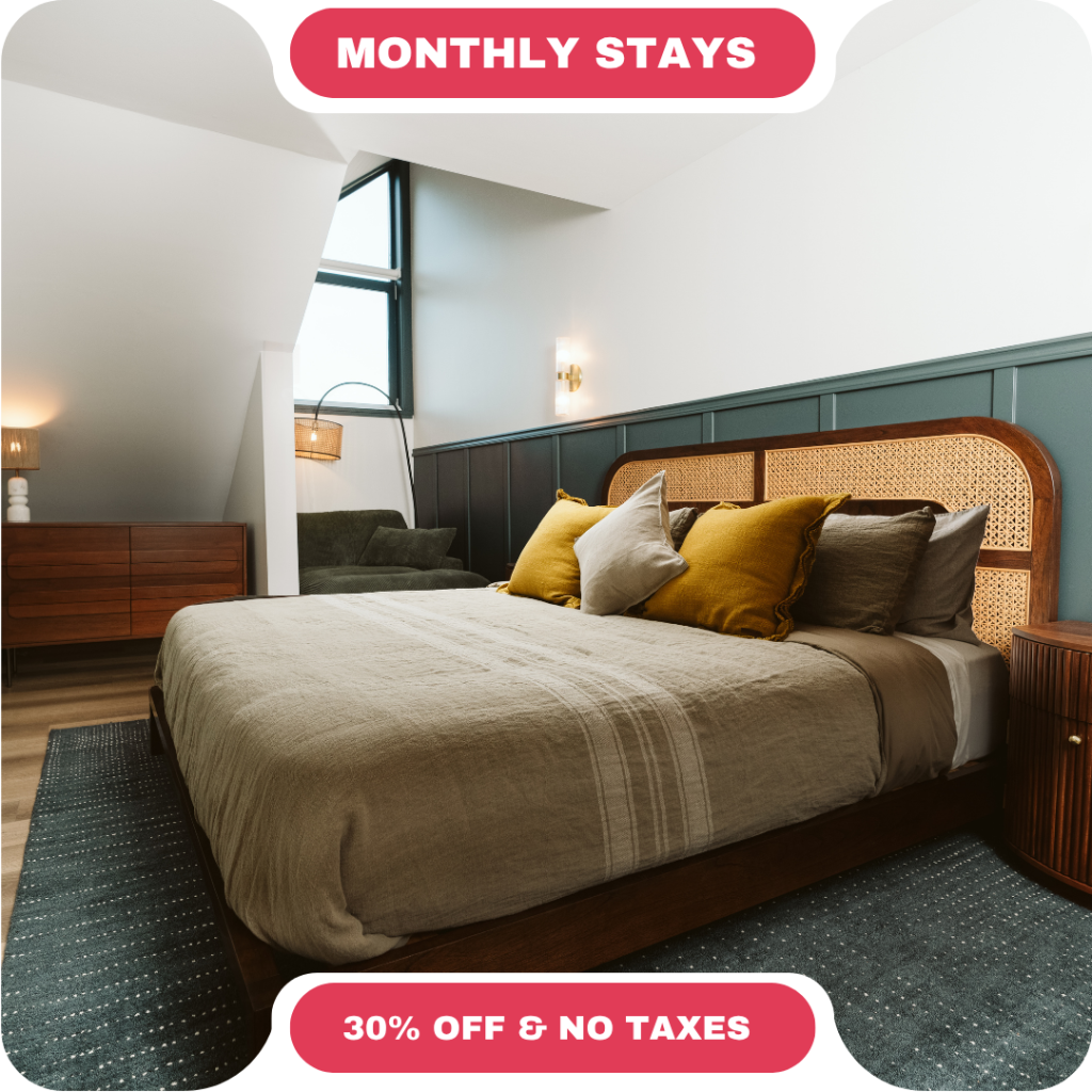 Modern bedroom with a large bed, decorative pillows, armchair, wooden furniture, and a large window—perfect for vacation rentals in Whistler. Text reads "Monthly Stays" and "30% Off & No Taxes.