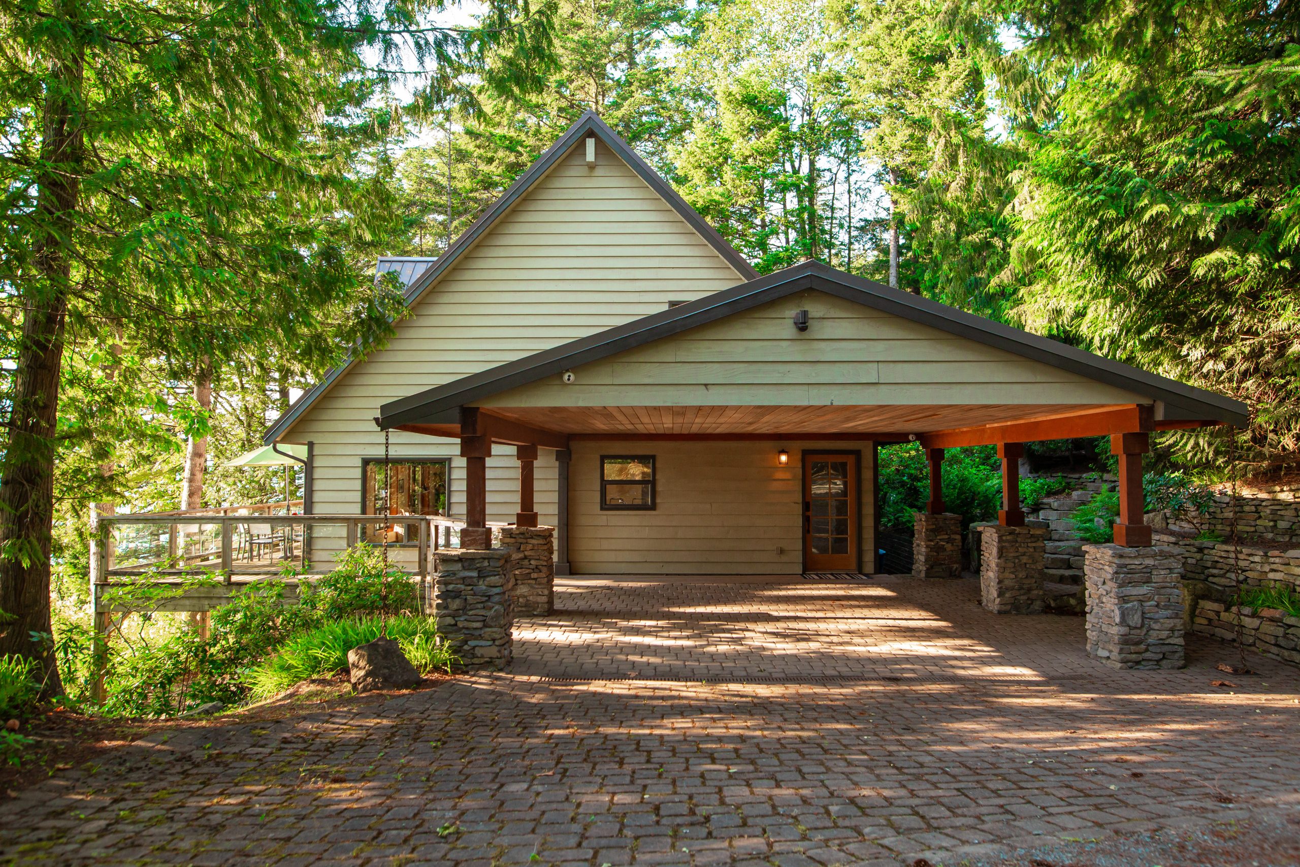 A beige house with a steep roof and covered carport evokes the apres vibe, surrounded by large trees and a stone-paved driveway.