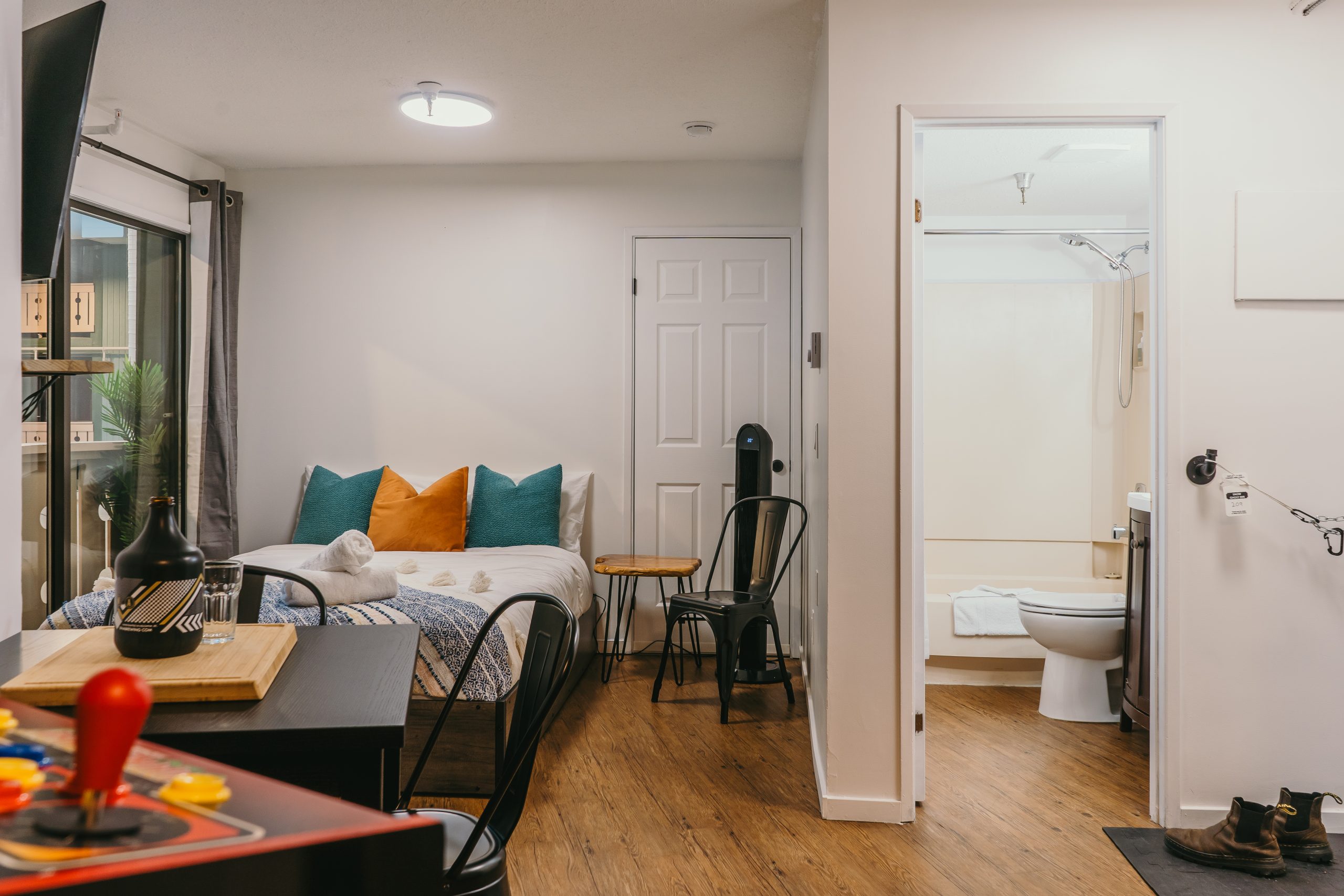 Studio apartment at The Apres featuring a bed, small dining table, chairs, an arcade machine, and an open door to a bathroom with accessible features.