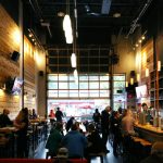 People sit at tables and the bar inside a modern, high-ceilinged brewery with wood-paneled walls, pendant lights, and large windows—one of the top Things to Do in Big White for those looking to relax with friends and craft beer.