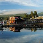 A wooden building and dock are reflected in calm water, with boats moored nearby—a serene scene that captures the natural beauty one can experience when buying property in Ucluelet, set against a forested, mountainous backdrop under a partly cloudy sky.