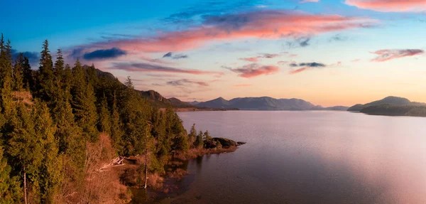 A calm lake bordered by dense evergreen forest under a colorful sunset sky with pink clouds and distant mountains—an idyllic scene reminiscent of buying property in Ucluelet.