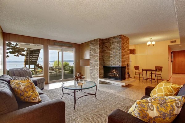 Spacious living room with two couches, glass coffee table, brick fireplace, dining area, and large windows overlooking an outdoor patio and stunning water views—perfect for those interested in buying property in Ucluelet.