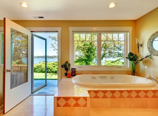 Spacious bathroom with a large bathtub, open door leading to a patio, and windows showcasing lush greenery and water—perfect for those buying property in Ucluelet seeking comfort and scenic views.