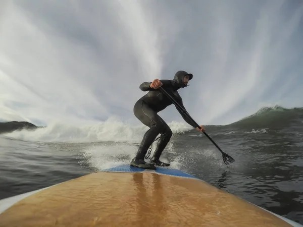 A person in a wetsuit paddleboards on a wave, balancing on a surfboard in the ocean under a cloudy sky—capturing the adventure that comes with buying property in Ucluelet.