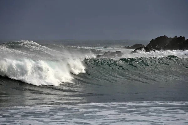 Large ocean waves crash near dark rocky outcrops under a cloudy gray sky, offering a breathtaking view perfect for those interested in buying property in Ucluelet.