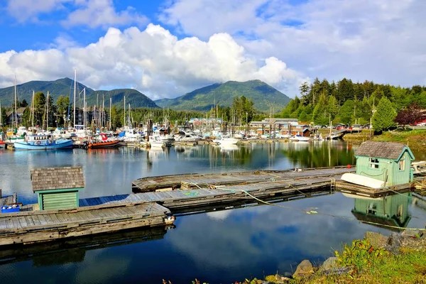 A harbor with boats docked at wooden piers, small green sheds, calm water reflecting the sky, and forested mountains in the background under partly cloudy skies—an inviting scene for those considering buying property in Ucluelet.