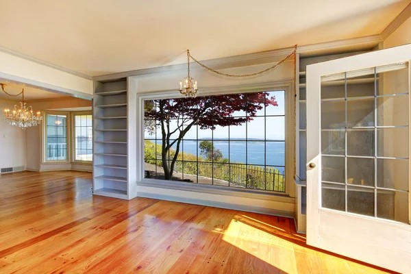 Bright room with wood floors, chandeliers, built-in shelves, and a large picture window. An open glass door reveals trees, railing, and water—perfect for anyone interested in buying property in Ucluelet.