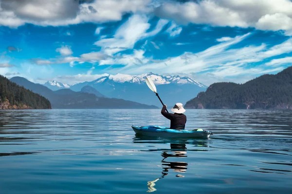 A person in a kayak paddles on a calm lake surrounded by mountains and a partly cloudy sky, enjoying one of the top fall activities Harrison Hot Springs has to offer.