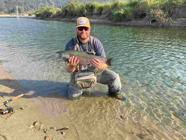 A person kneels in shallow river water at Harrison Hot Springs, holding a large fish with both hands. Wearing a hat, sunglasses, and fishing gear, they enjoy one of the top fall activities as trees and hills complete the scenic backdrop.