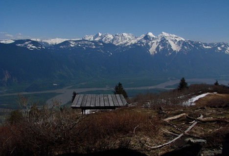 Wooden bench on a mountain overlook near Harrison Hot Springs, with snow-capped peaks and a river valley in the background—perfect for Fall activities under a clear blue sky.
