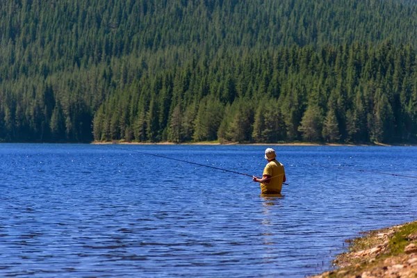 A person wearing a yellow shirt and hat stands in a lake, fishing with a rod—one of the relaxing spring activities in Harrison Hot Springs. Dense green forest and hills form a scenic backdrop.