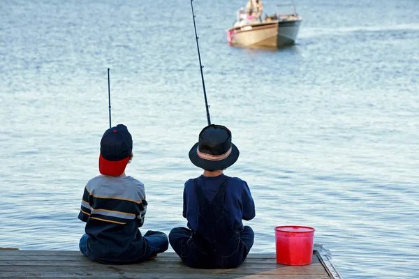 Two children sit on a dock fishing with rods, facing the lake at Harrison Hot Springs. A red bucket rests beside them as a boat drifts in the background—perfect for spring activities and fun things to do outdoors.