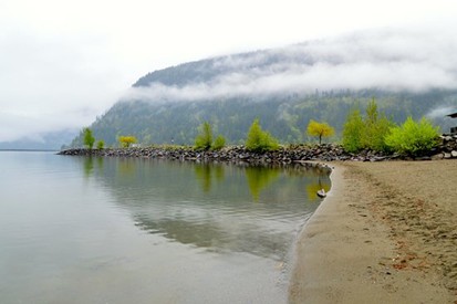 A calm lake with a sandy shore and rocky edge lined by green bushes, set near tree-covered hills draped in low clouds—perfect for relaxing or exploring spring activities in Harrison Hot Springs.