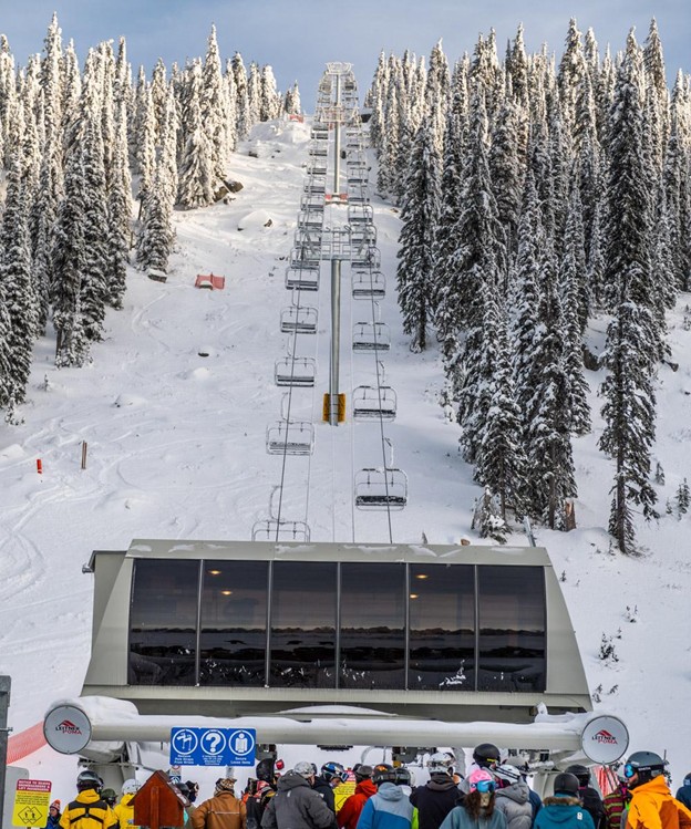 Skiers and snowboarders wait in line at the base of a ski lift on a snowy mountain, with pine trees and empty chairlifts ascending the slope—hinting at the excitement surrounding Big White expansion plans.
