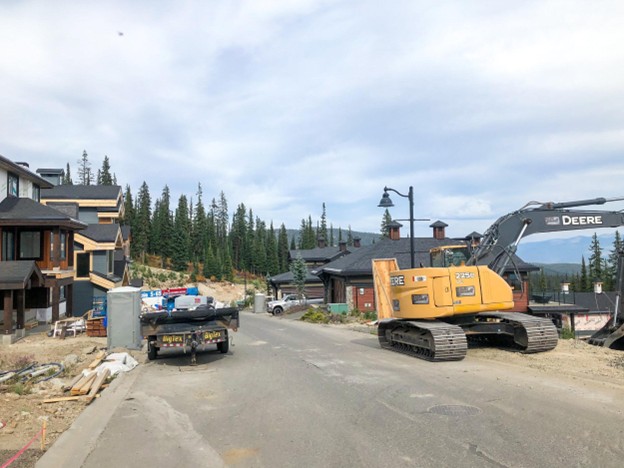 Residential neighborhood under construction with a yellow excavator, a truck, and building materials on the street; modern houses and pine trees in the background reflect the area's Big White expansion plans.