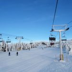 Skiers and snowboarders move across a snowy slope under a blue sky, with multiple ski lifts carrying people above the scene—an atmosphere buzzing with excitement about Big White expansion plans. Snow-covered trees surround the area.