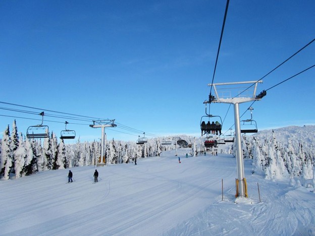 Skiers and snowboarders move across a snowy slope under a blue sky, with multiple ski lifts carrying people above the scene—an atmosphere buzzing with excitement about Big White expansion plans. Snow-covered trees surround the area.