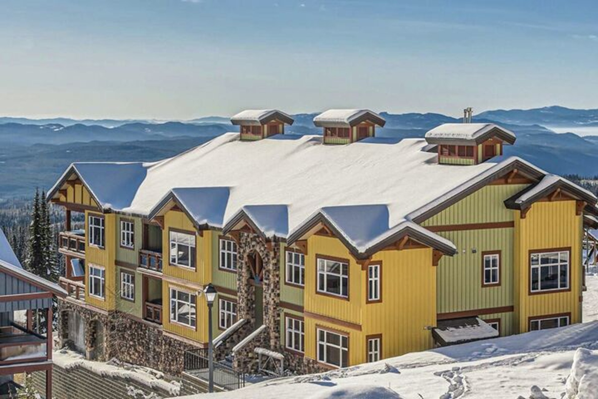 A large yellow alpine-style lodge with a snow-covered roof sits on a snowy hillside, hinting at Big White expansion plans amid distant mountains and a clear blue sky.