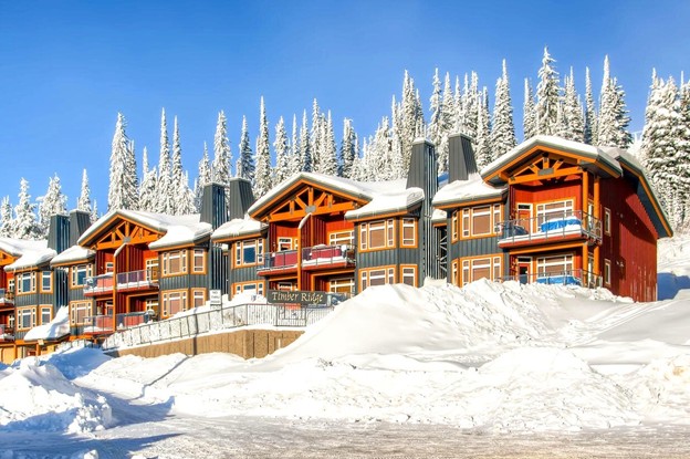 A row of colorful, chalet-style lodges with snow-covered roofs sits beneath tall evergreen trees under a clear blue sky, hinting at the charm promised by upcoming Big White expansion plans.