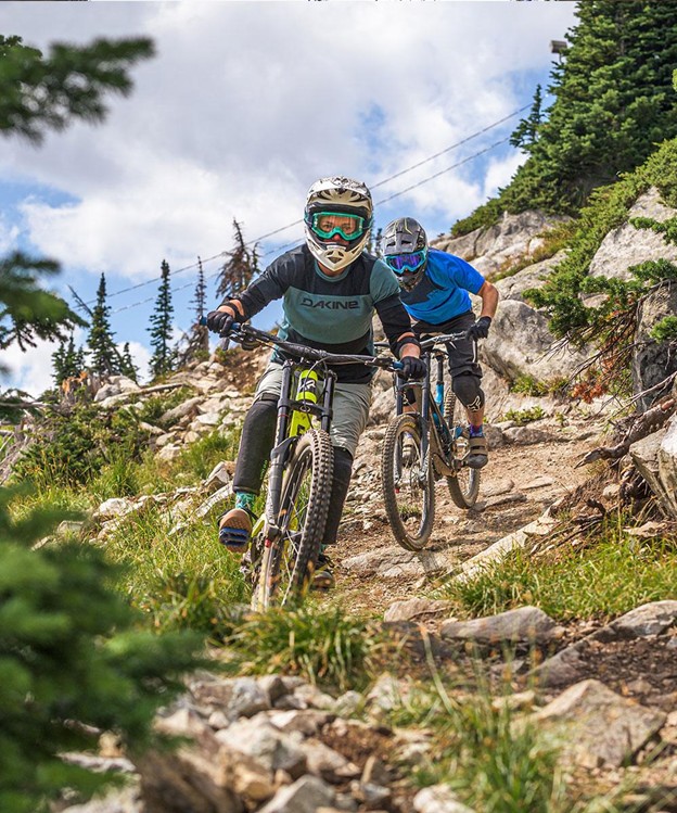 Two mountain bikers wearing helmets and goggles ride down a rocky dirt trail surrounded by trees and shrubs on a partly cloudy day, exploring new paths inspired by Big White expansion plans.