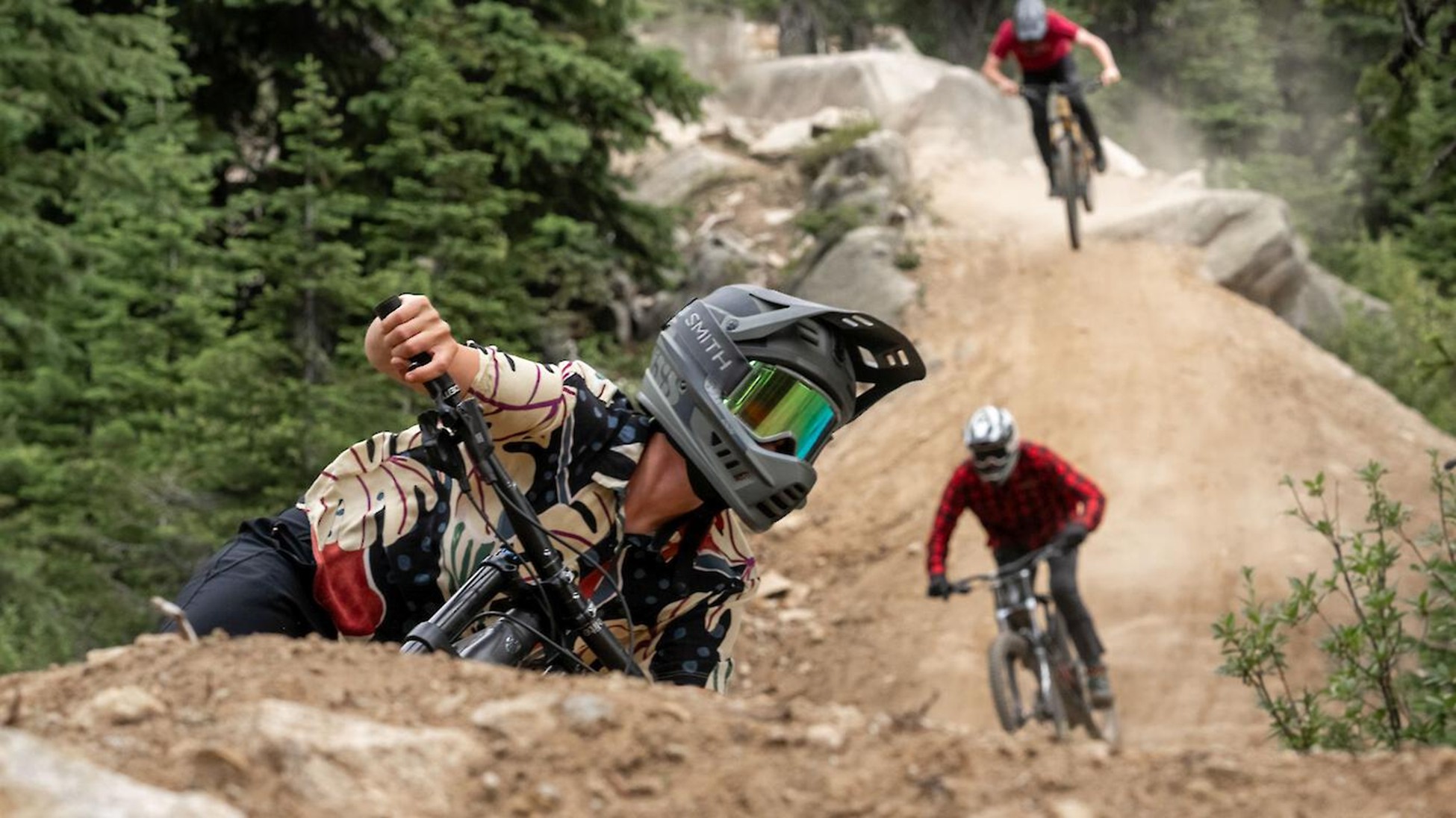 Three mountain bikers ride down a dirt trail in a forest at Big White; the closest rider leans into a turn while the others follow behind, one catching air over a jump—hinting at excitement for Big White expansion plans.