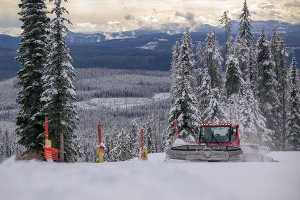 A red snow groomer drives on a snowy slope surrounded by tall pine trees, with mountains in the background—hinting at Big White expansion plans on the horizon.