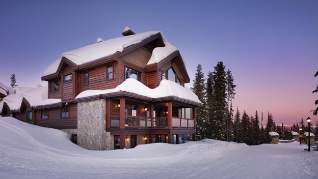 A large wooden cabin covered in snow sits among pine trees at dusk, surrounded by snowbanks and a clear sky—a scene reminiscent of Big White expansion plans in this tranquil winter landscape.