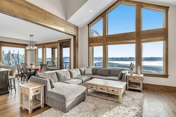 Modern living room with a large gray sectional sofa, wooden tables, and floor-to-ceiling windows showcasing mountain and lake views—perfect for relaxing after exploring the nearby Big White expansion plans. Dining area visible in the background.