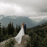A bride and groom stand on a large rock surrounded by pine trees, mountains, and clouds in the background—capturing the magic of a Wedding in Whistler.