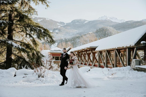 A bride and groom walk together outside in the snow near a wooden bridge, surrounded by trees and mountains—capturing the magic of planning a wedding in Whistler.