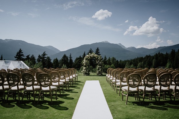 Rows of empty chairs face a floral arch on a grassy outdoor aisle, with mountains and trees in the background under a partly cloudy sky—perfect inspiration for planning a wedding in Whistler.